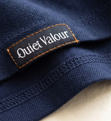 Close-up of a navy blue textured fabric garment with a black brand label reading 'Quiet Valour' in white text, bordered by orange stitching.