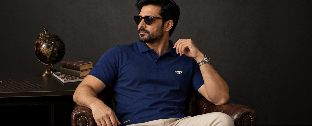 A man wearing a navy blue polo shirt with gold embroidered text on the chest, styled with dark sunglasses and a silver watch, seated in a leather armchair against a dark gray background.slider_item_Qaethp
