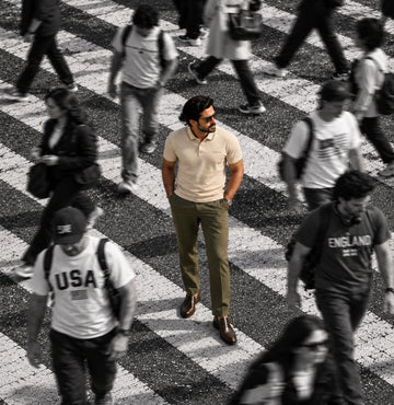 A man wearing a light beige polo shirt, olive green trousers, and brown shoes walks across a black-and-white striped crosswalk, with other pedestrians blurred in the background.