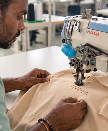 A close-up lifestyle shot of a man operating a Pegasus industrial sewing machine in a workshop, stitching a beige t-shirt with 'INDIA' embroidered on it.