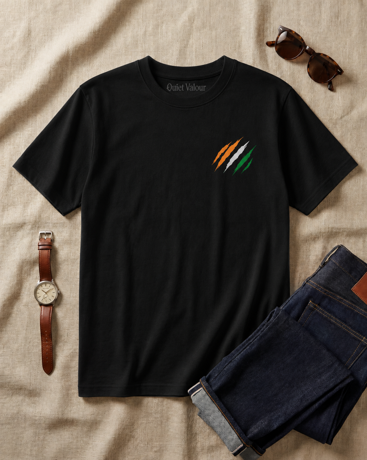 A black short-sleeve t-shirt with a small graphic of three diagonal stripes in orange, white, and green on the left chest, styled in a flat lay on a beige linen background with dark blue jeans, sunglasses, and a brown leather watch.