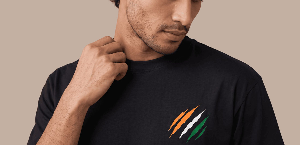 A close-up of a man wearing a black t-shirt with a graphic of three diagonal stripes in the colors of the Indian flag (orange, white, and green) on the left chest, set against a solid beige background.