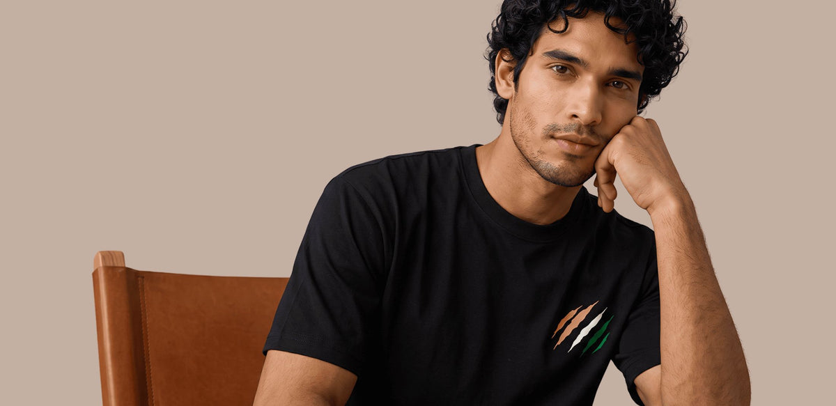 A man wearing a black short-sleeve t-shirt with a small embroidered graphic on the left chest, sitting in a brown leather chair against a solid beige background.