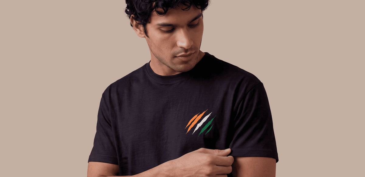 A man wearing a black t-shirt with a small graphic of three diagonal stripes in saffron, white, and green on the left chest, set against a solid beige background.