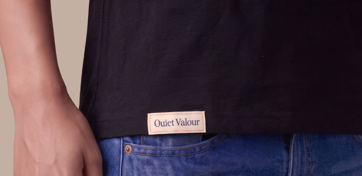 Close-up of a person wearing a black t-shirt with a beige 'Quiet Valour' brand patch on the front, paired with blue jeans.