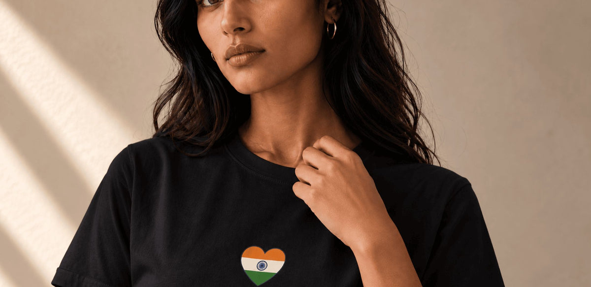 A woman wearing a black crewneck t-shirt with a small heart-shaped Indian flag graphic on the chest, set against a warm beige background with soft diagonal shadows.