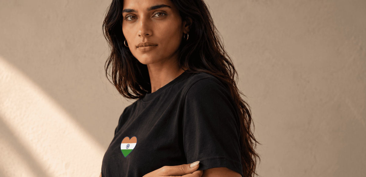 A woman with long dark hair wearing a black t-shirt with a small heart-shaped Indian flag graphic on the left chest, posed against a textured beige wall with soft window light casting shadows.