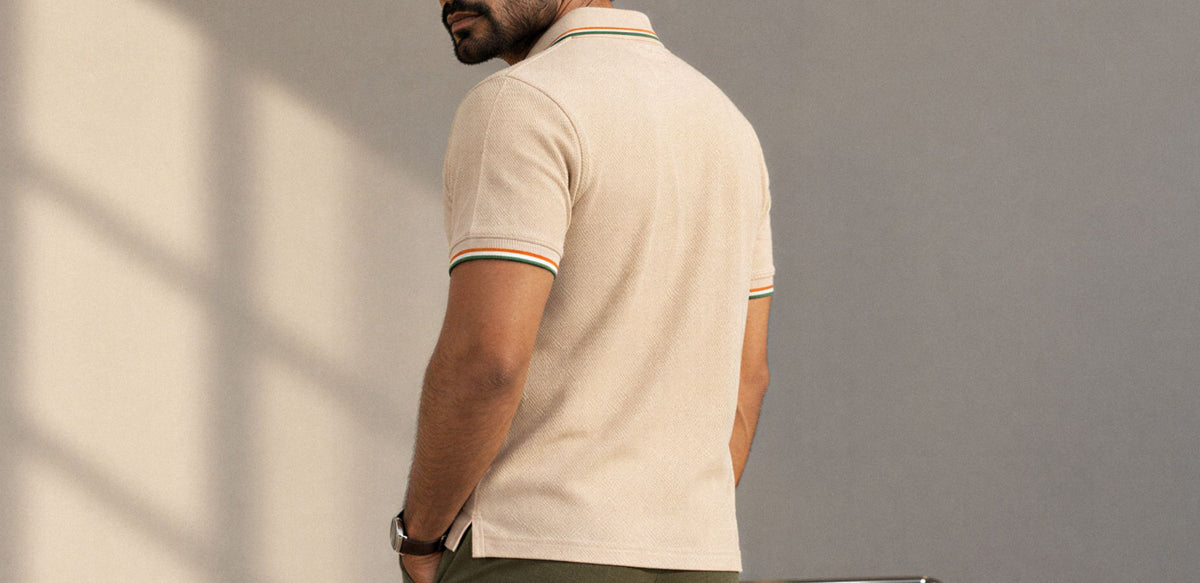 A man shown from the back wearing a cream-colored short-sleeve polo shirt with thin green and orange stripes on the collar and sleeve cuffs, set against a gray wall with soft window shadows.