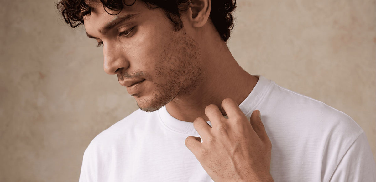 A man with curly dark hair and light stubble, wearing a plain white crew-neck t-shirt, looking down and touching his neck against a textured beige background.