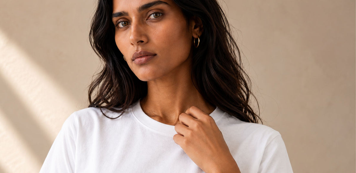 A woman with long dark hair wearing a plain white crew-neck t-shirt, shown from the chest up against a warm beige background.