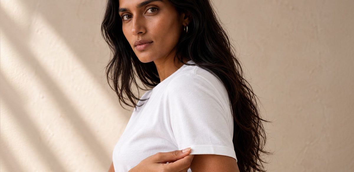 A woman with long dark hair wearing a plain white short-sleeve crewneck t-shirt, shown from the chest up against a textured beige wall with soft diagonal shadows.